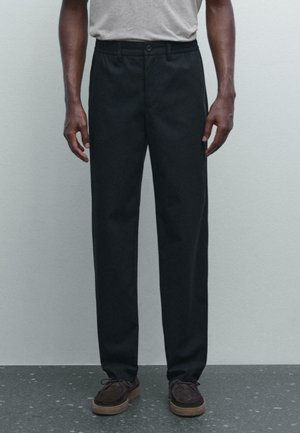 Pantalon noir, coupe droite, avec une taille plate, en mélange de coton, poches latérales et fermeture à bouton. Porté avec des chaussures marron.