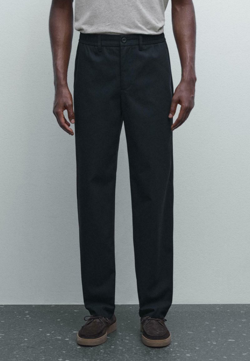 Black, straight-fit trousers with a flat front, cotton blend material, side pockets, and a button closure. Worn with brown shoes.