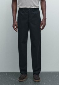 Man wearing black straight-leg trousers, gray tucked-in shirt, and brown lace-up shoes standing on speckled floor against plain wall.