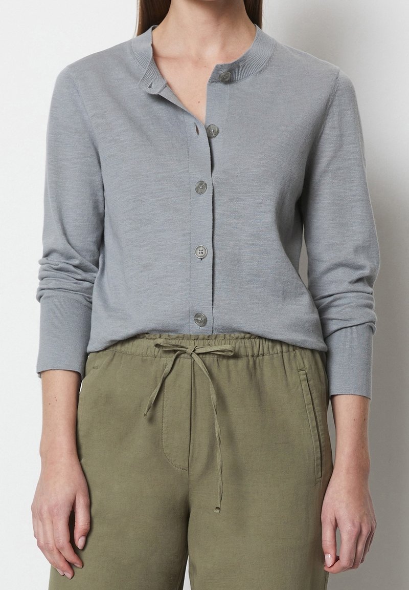 Femme portant un cardigan boutonné gris clair avec un col rond et un pantalon vert olive à cordon, debout devant un fond blanc.
