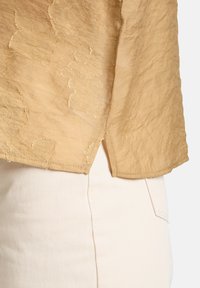 Top a maniche corte di un leggero beige con un motivo floreale testurizzato, con orlo grezzo e spacchi laterali, abbinato a jeans chiari.