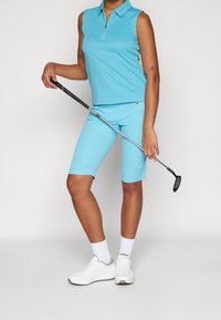 Polo sin mangas azul claro con cierre de cremallera, combinado con pantalones cortos a juego hasta la rodilla. Zapatillas deportivas blancas y un palo de golf.