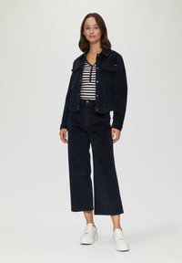 Marineblauwe corduroy jas en bijpassende wijde broek gecombineerd met een gestreept shirt. Het model draagt witte sneakers en toont een casual outfit.