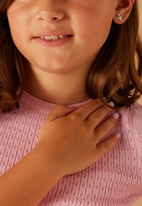 Roze geribbeld gebreid topje met een ronde halslijn, met een hand die op de borst rust. Kleine zilveren studoorbel met een kleurrijk ontwerp.