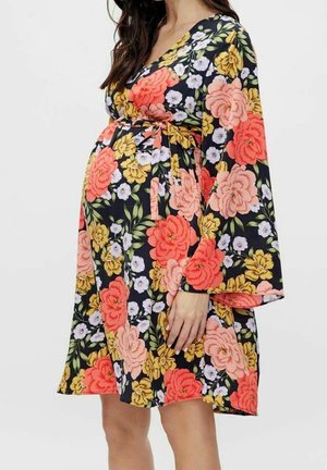 Gravid kvinde iført en sort blomsterkjole med pink, orange, gule og hvide blomster og vide ærmer, stående med hånden hvilende på maven.