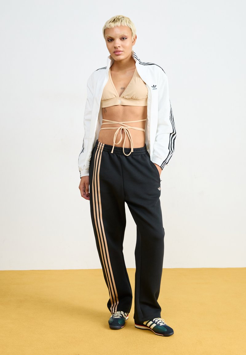 Pantalones de chándal negros con rayas doradas, un top corto beige con lazos y una chaqueta blanca con rayas negras, combinados con zapatillas coloridas.