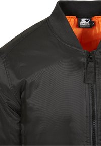 Giubbotto bomber nero con una texture liscia e lucida, chiusura con zip e un vivace rivestimento interno arancione. Presenta un colletto a coste.
