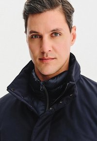 Granatowa kurtka puchowa z wysokim kołnierzem, z zamkiem z przodu i zatrzaskami. Materiał wydaje się gładki i błyszczący z wypukłą teksturą.