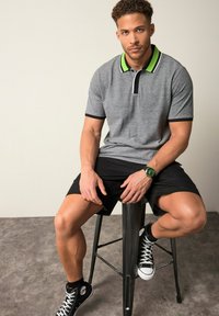 Grå polo t-shirt med grön och svart randig krage, tillsammans med svarta shorts. Modell som bär en grön klocka och svarta sneakers, sittande på en pall.
