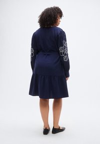 Φόρεμα στο χρώμα του ναυτικού μπλε με φουσκωτά μανίκια, κεντητές ανθικές λεπτομέρειες στα μανίκια, εφαρμοστή μέση και βολάν στο κάτω μέρος. Συνδυάζεται με μαύρα φλατ παπούτσια.