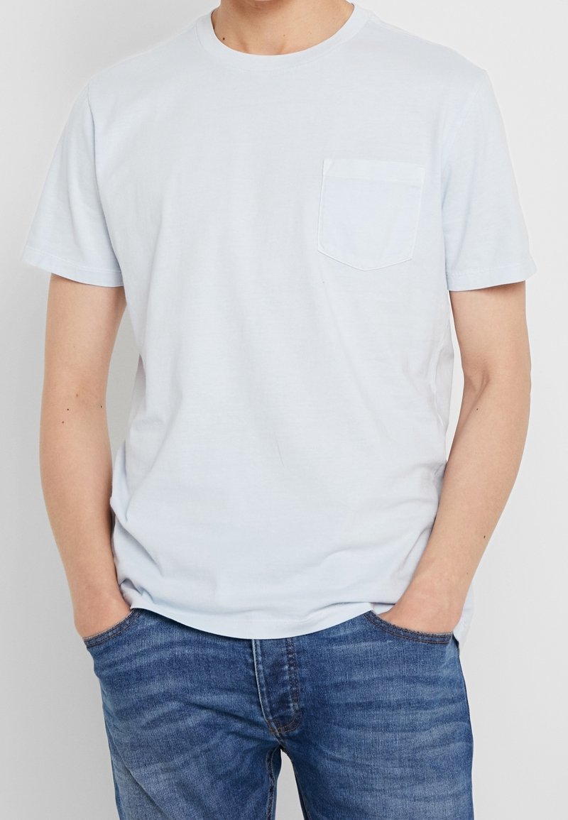 T-shirt en coton bleu clair avec un col rond et des manches courtes. Présente une poche sur la poitrine gauche et une texture douce et lisse.