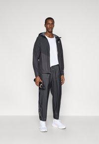 Μαύρο και γκρι μπουφάν με κουκούλα, συνδυασμένο με ταιριασμένα φαρδιά παντελόνια και λευκά αθλητικά παπούτσια. Το μοντέλο κρατάει μια μαύρη φιάλη νερού.