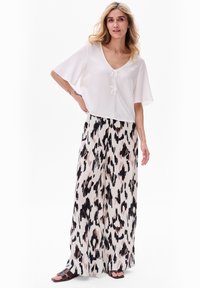 Biała bluzka z krótkim rękawem, dekoltem w kształcie litery V i detalem w postaci wiązania, zestawiona z szerokimi spodniami w czarno-beżowy wzór w panterkę. Widoczne płaskie sandały.