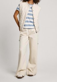 Béžová prošívaná vesta přes modro-bíle pruhované tričko, doplněná světle béžovými kalhotami s širokými nohavicemi a jednoduchými šedými botami.
