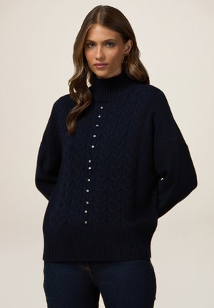 Maglione lavorato a maglia blu navy con collo alto, caratterizzato da un design a trecce e bottoni decorativi sul davanti. Texture morbida e vestibilità comoda.