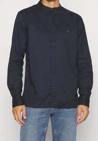 Koszula z długim rękawem w kolorze granatowym, z kołnierzykiem typu band, z przodem na guziki i subtelnym logo na klatce piersiowej. Tkanina wydaje się gładka i ma luźny krój.
