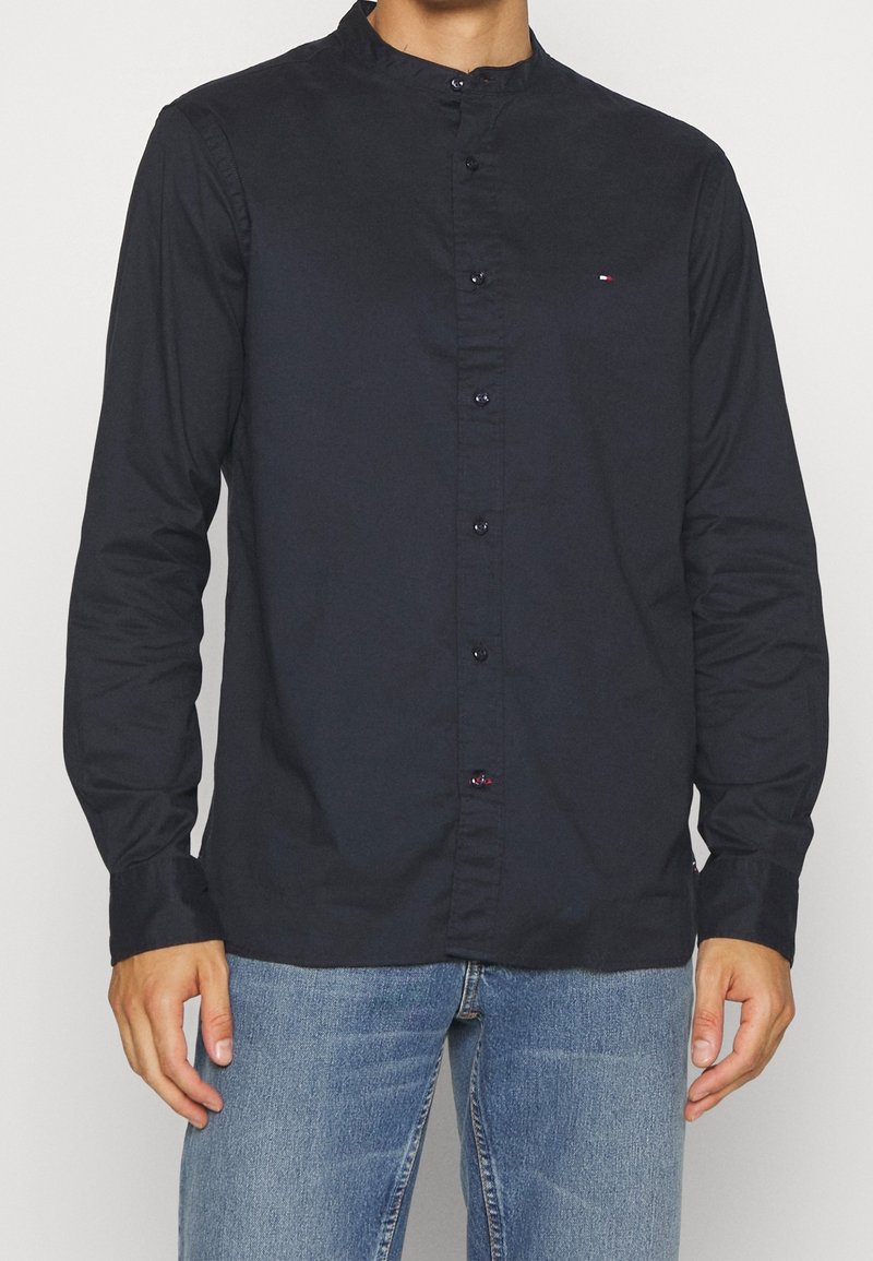 Koszula z długim rękawem w kolorze granatowym, z kołnierzykiem typu band, z przodem na guziki i subtelnym logo na klatce piersiowej. Tkanina wydaje się gładka i ma luźny krój.