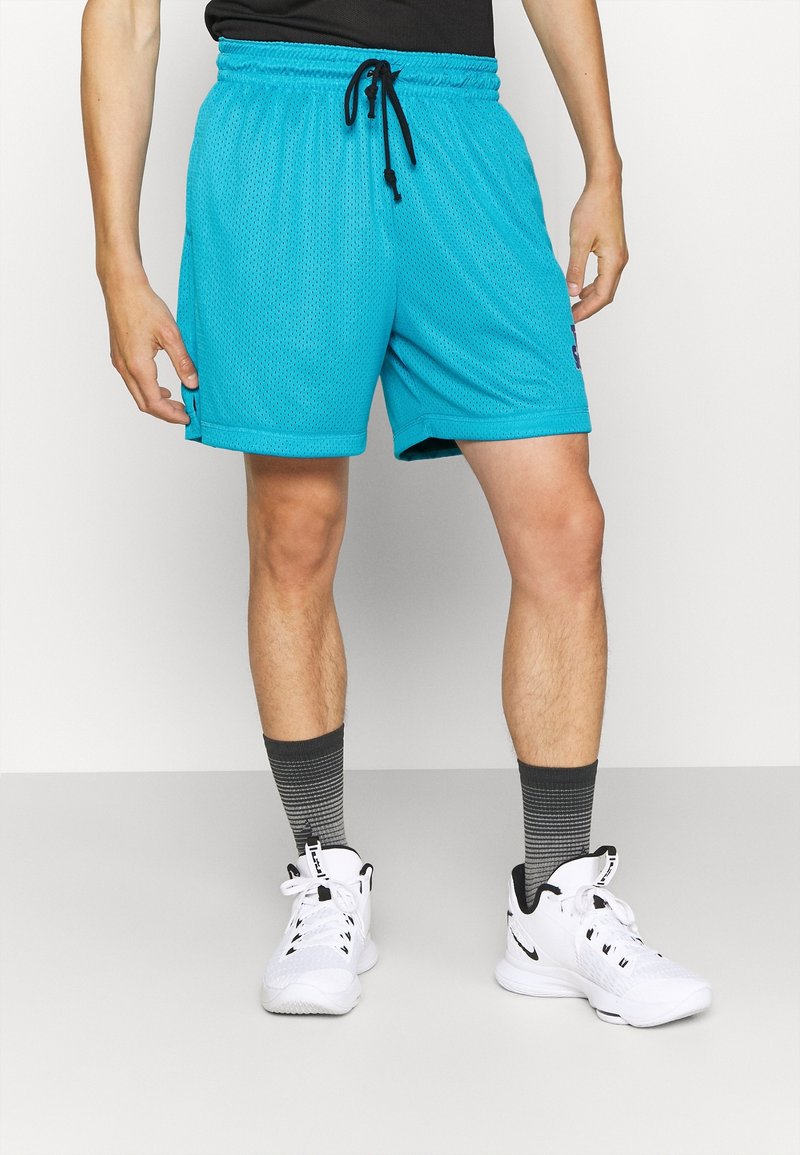 Homme portant un short de sport en mesh turquoise, des chaussettes montantes noires dégradées et des baskets blanches montantes, debout devant un fond blanc uni.
