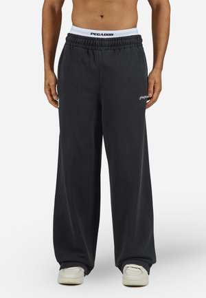 Black, loose-fit sweatpants with an elastic waistband featuring white branding. Fabric appears soft, with a smooth texture and no patterns.