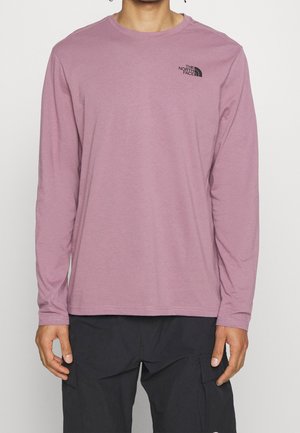 Mann trägt ein mauvefarbenes Langarmshirt mit dem The North Face-Logo auf der linken Brust und schwarze Cargohosen.