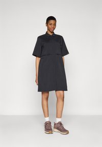 Vestito nero a maniche corte con colletto, dettagli con zip in vita, abbinato a scarpe da ginnastica marroni e grigie e calzini bianchi.