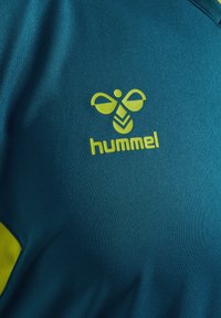 Teal sportströja med en strukturerad tygkvalitet, som har en gul Hummel-logotyp och en abstrakt ikon, kompletterad med en gul accent på sidan.