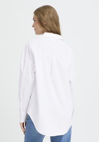 White button-up shirt with vertical stripes, long sleeves, and a back pleat; paired with light blue jeans and a smooth fabric texture.