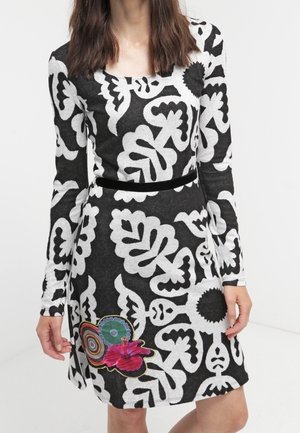Woman wearing a long-sleeve black and white patterned dress with a colorful floral and circular patch near the hem.
