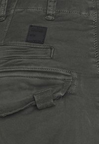 Donkergroene broek met een gestructureerde stof, meerdere zakken en een zwart label met "Alpha Industries" in reliëf. Subtiele stikseldetails zichtbaar.