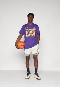 Violetinė marškinėliai su "Los Angeles Lakers" geltonai, balti šortai su juodais akcentais, laikantis krepšinio, vilkint juodus ir auksinius sportbačius.