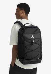 Black backpack featuring a textured finish, adjustable straps, and a distinctive bungee cord design on the front. Includes side pockets.