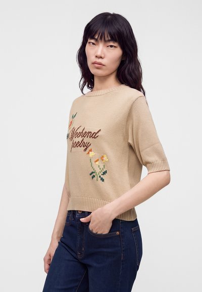 Femme aux longs cheveux foncés portant un pull beige à manches courtes avec le texte "Weekend poetry" et une broderie florale, ainsi qu'un jean bleu foncé.