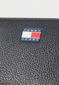Black leather wallet with a textured surface, featuring a rectangular Tommy Jeans logo in blue, red, and white on the front.