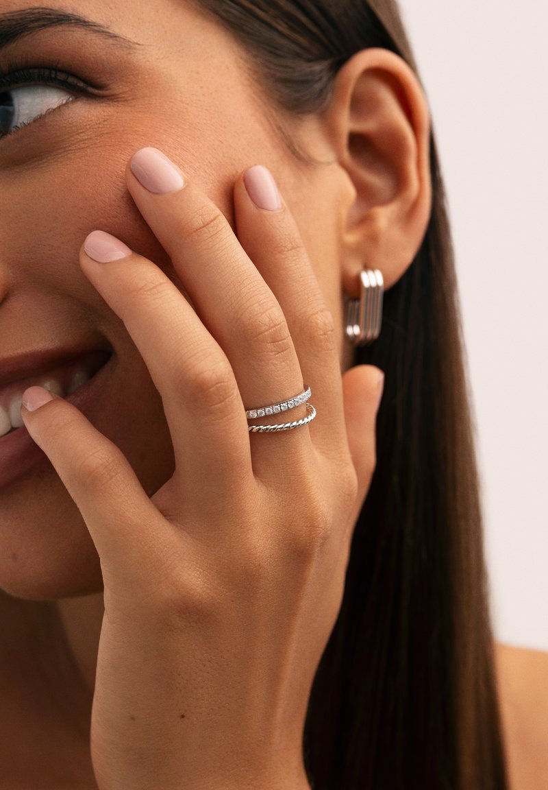 Mujer sonriendo con la mano tocando la mejilla, que lleva anillos plateados retorcidos con acentos de diamantes y pendientes de aro plateados a rayas.