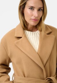 Cappotto cammello in tessuto testurizzato, con ampie rever e una cintura annodata in vita, indossato sopra un maglione a maglia color crema.