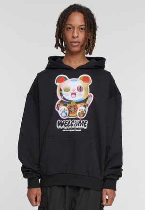 Zwarte hoodie met een kleurrijke afbeelding van een gelukskat en de tekst "WELKOM GOEDE FORTUNE." Losse pasvorm met een kangoeroezak.