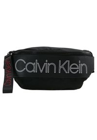 Le sac pochette noir Calvin Klein présente un extérieur en nylon lisse, un logo blanc audacieux, une fermeture éclair et une sangle en tissu avec un accent de logo.