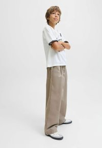 Ragazzo con capelli castano chiaro sta in piedi con le braccia incrociate indossando una maglietta bianca con grafica, pantaloni beige larghi e sneakers bianche su uno sfondo bianco.