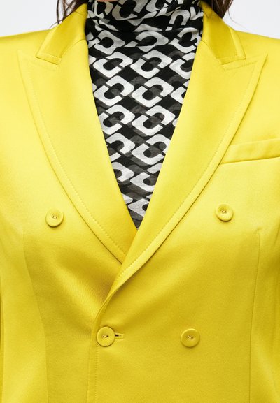 Blazer giallo doppiopetto con rever a incavo e due bottoni, indossato sopra un dolcevita nero e bianco con motivi geometrici.