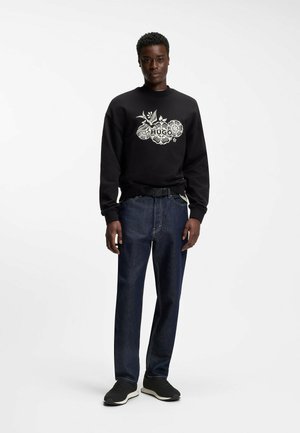 Mann steht, trägt schwarzen Sweatshirt mit floralem Muster, dunkelblaue Jeans, schwarzen Gürtel und schwarze Turnschuhe vor weißem Hintergrund.