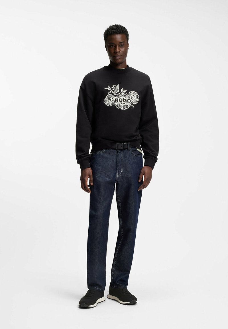 Homme debout portant un sweat-shirt noir à motif floral, un jean bleu foncé, une ceinture noire et des baskets noires sur fond blanc.
