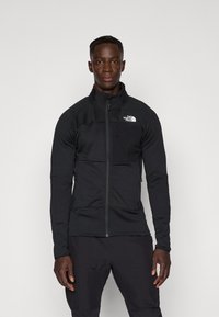 Svart zip-up jakke med høy krage, teksturert stoff og brystlomme. Logo på venstre skulder. Egnet for utendørs bruk.