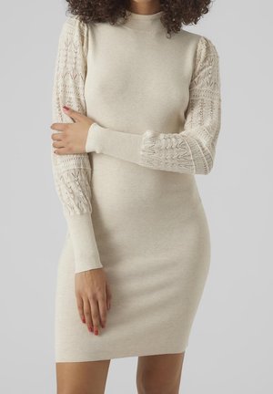 Kvinde iført beige, figursyet strikkjole med mønstrede ærmer i fuld længde, stående med en arm på tværs og afslappet kropsholdning mod en ensfarvet baggrund.