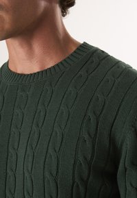 Zielony sweter dzianinowy z splotem warkoczowym i prążkowanym okrągłym dekoltem. Zbliżenie ukazujące teksturę i detale tkaniny.