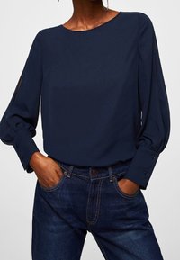 Blouse bleu marine à manches longues avec un col rond, des manches bouffantes et des poignets boutonnés, accompagnée d'un jean bleu foncé. Texture de tissu lisse.