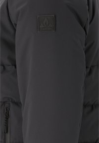 Schwarze gepolsterte Jacke aus strukturiertem Stoff, mit einem Reißverschlussfach und einem Gummilogo-Patch auf der linken Schulter.