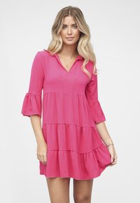 Vaaleanpunainen kerroksittain laskostettu mekko, jossa on kolme neljäsosaa hihoista kellomalliset hihat, V-aukko ja teksturoitu kangas, mekko on polven yläpuolelle yltävä.