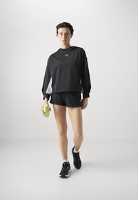 Sudadera negra de gran tamaño con capucha, combinada con pantalones cortos texturizados negros. Zapatillas negras con acentos verdes. Sostenido una botella de agua verde claro.