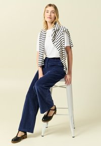 Marinblå kordflöjt vida byxor i kombination med en vit t-shirt med rund halsringning, accentuerad av en marinblå och vit randig sjal runt axlarna. Svarta klackar.