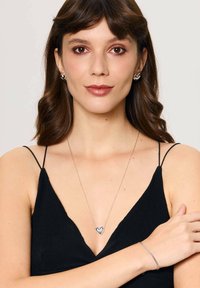 Srebrny zestaw biżuterii, w tym naszyjnik z serduszkowym wisiorkiem oraz pasujące kolczyki. Modelka ma na sobie czarny top na ramiączkach.